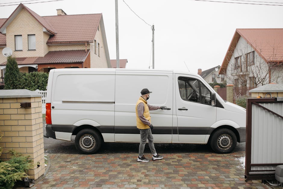 A man wearing a face mask, a yellow and gray jacket, and dark trousers is standing outside a residential property next to a white panel van, which is parked on a paved driveway. The man is holding the van's sliding door handle, preparing to load or unload items during a house removal process. The van is positioned parallel to the house, with a large open door visible on the side near the man, indicating active loading or unloading of furniture, boxes, or packing materials. The environment includes neighboring houses with brick and weatherboard exteriors, red tiled roofs, and front gardens enclosed by low brick walls and fences. The scene takes place in daylight, with overcast skies suggesting typical weather conditions for home relocation and furniture transport in North Harrow. The presence of packing supplies such as cardboard boxes, plastic wrapping, or blankets is not visible in this specific image, but the context aligns with packing and moving services provided by Man with Van North Harrow as part of professional house removals.