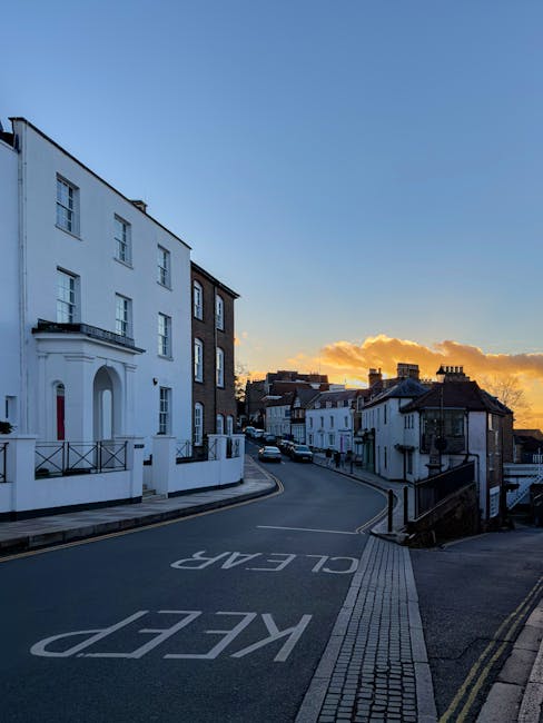 Photograph of a quiet residential street during sunset, featuring a white multi-storey building with large windows and a balcony on the left, adjacent to a darker brick building. The street is gently curved, with a designated 'KEEP CLEAR' area painted on the asphalt, and a tactile paving strip along the sidewalk on the right side. In the distance, several rows of terraced houses with pitched roofs extend along the road. The sky displays a mix of blue and warm orange hues with a few clouds illuminated by the setting sun. The scene suggests an urban environment suitable for home relocations and furniture transport services, with no vehicles or moving activity visible at this moment. This image could be associated with professional removals in North Harrow, highlighting the residential setting where house moves and packing processes occur, with Man with Van North Harrow providing the relevant services.