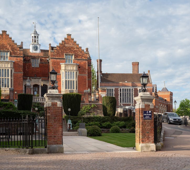 A view of a historic brick building with a clock tower and ornate architectural details, situated behind a gated entrance with brick pillars topped by black iron lanterns. In the foreground, a paved pathway leads from the street to the building, bordered by well-maintained grass and trimmed hedges. The scene includes a vehicle parked near the entrance and a flagpole extending upward from the property. This setting reflects a location suitable for residential or official functions, with the surrounding area indicating potential for house removals and furniture transport. Man with Van North Harrow may coordinate home relocation services in such environments, involving careful packing, loading, and transport of household furniture and boxes visible along the pathway and driveway.