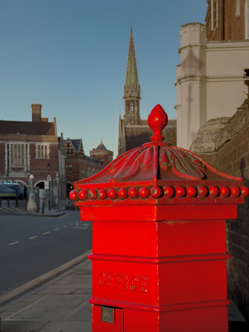 A close-up view of a bright red traditional UK postal box situated on a sidewalk during daylight hours, with the word 'OFFICE' embossed on its front. Behind the postal box, there is a stone building with decorative architectural details and a parapet wall. In the background, a church spire with a pointed roof and gothic features rises against a clear blue sky, indicating a historic part of North Harrow. To the left, parked cars line the street, and a few pedestrians are visible walking along the pavement. The scene depicts an urban environment typical of North Harrow, relevant to house removals and local logistics, with a focus on the street setting where furniture transport or packing and moving activities might take place, especially in relation to complying with local council permits for home relocation, as referenced in the page title.