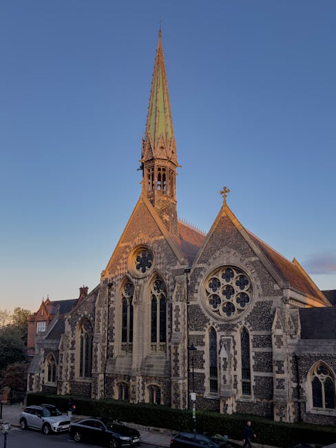A detailed exterior view of a historic church with intricate stonework and large stained glass windows featuring circular and pointed arch designs. The church has a tall, pointed spire with a greenish patina, adorned with a small cross at the top, rising above the building. The stone walls display a mixture of light and dark patches, indicating weathering and age, with lancet windows and decorative stone motifs. In the foreground, several cars are parked along the street, and a few pedestrians are visible walking nearby on the pavement. The church is situated in a residential or urban area, with some houses and trees in the background under a clear evening sky. This image is related to house removals and moving services, highlighting the type of landmark near Harrow on the Hill, consistent with the content about home relocation and furniture transport services offered by Man with Van North Harrow.