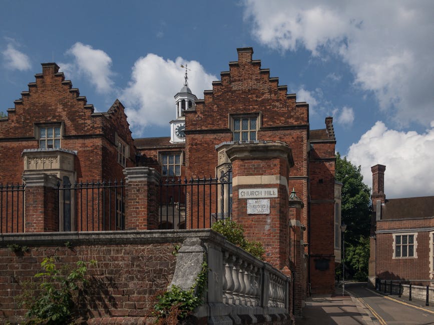 The image depicts a historic red brick building with a steep gabled roof, decorative stone window surrounds, and a small clock tower at the top, situated behind a wrought iron fence on a sidewalk. The building has several large windows and features a stone sign that reads 'Chuch Hill' on a brick pillar in front of the fence, indicating the location. The sky above is partly cloudy with patches of blue, and there are some green trees visible behind the building. The scene captures the exterior of a heritage site characteristic of traditional British architecture, possibly a church or historic institution, with a nearby smaller brick building on the right, and a paved street with road markings running in front. This setting aligns with the context of house removals and relocation services, which often involve coordinating transport near such landmarks.