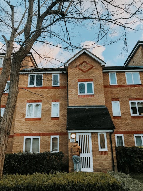 A man wearing a brown jacket and grey pants standing outside a three-storey residential building, facing a white door with vertical slats beneath a small black tiled awning, likely preparing for home relocation or a furniture transport service. The building features a brick facade with red brick accents around the windows, and the windows are white-framed with multiple panes. The scene is set during daylight, with a tall leafless tree in the foreground and some shrubbery along the base of the building. The sky is partly cloudy, providing natural light to the scene. This setup reflects typical logistics involved in house removals or packing and moving services provided by companies such as Man with Van North Harrow.