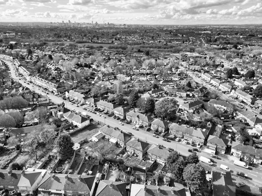 Black and white aerial photograph of a suburban residential area showing a long, straight street lined with terraced and semi-detached houses, with parked cars on both sides. In the foreground, rooftops and gardens of individual homes are visible, some with trees and fencing. The street extends into the distance, leading towards a more developed urban skyline with skyscrapers visible on the horizon. The scene captures a typical home relocation environment, where a house removal service such as Man with Van North Harrow might be involved in coordinating furniture transport, packing, or loading processes within this densely built neighbourhood.