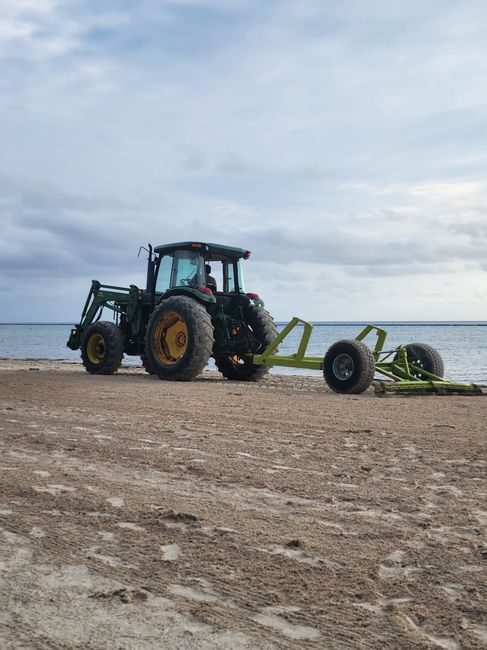 A green tractor with yellow wheels is parked on a sandy beach near the shoreline, positioned sideways to the camera. Attached to the tractor is a large, green agricultural implement with four wheels, lying flat on the ground and partially extended. The beach features a flat, textured surface with tire tracks and scattered small pebbles. The background shows a calm sea under a cloudy sky with soft, diffused lighting, and the horizon line separating water from the sky. The scene captures a rural or coastal setting, but in the context of home relocation and furniture transport, it exemplifies the use of machinery in logistical or outdoor moving operations, consistent with services offered by Man with Van North Harrow.