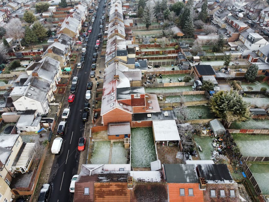 Black and white aerial photograph of a suburban residential area showing a long, straight street lined with terraced and semi-detached houses, with parked cars on both sides. In the foreground, rooftops and gardens of individual homes are visible, some with trees and fencing. The street extends into the distance, leading towards a more developed urban skyline with skyscrapers visible on the horizon. The scene captures a typical home relocation environment, where a house removal service such as Man with Van North Harrow might be involved in coordinating furniture transport, packing, or loading processes within this densely built neighbourhood.